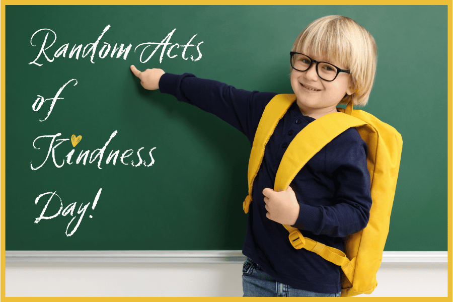 Young child with a yellow backpack standing in front of a chalkboard, pointing to the message "Random Acts of Kindness Day.