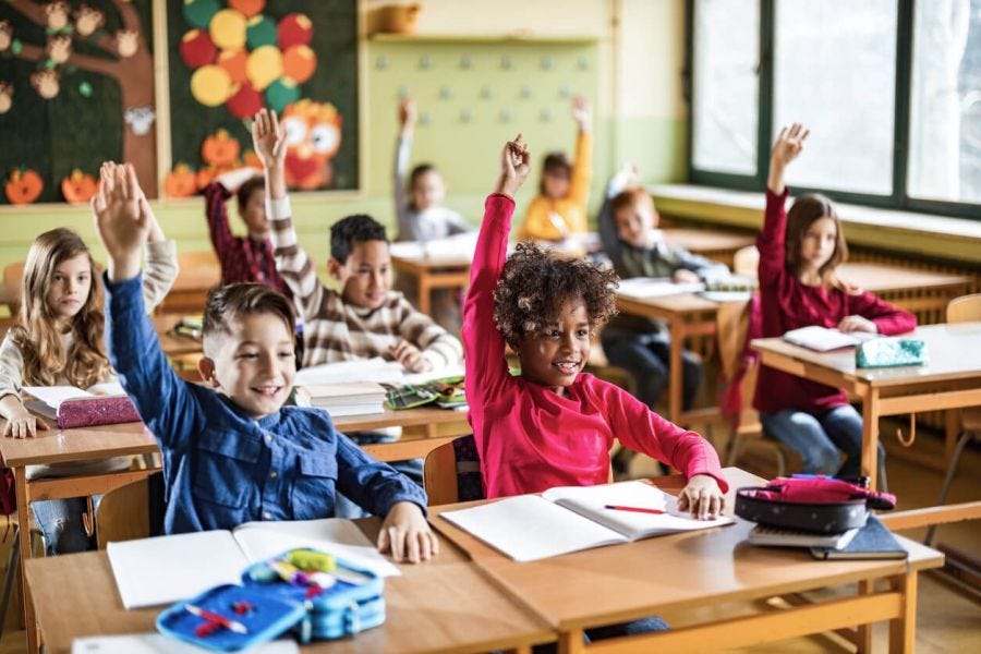 School classroom full of students sitting at desks raising their hands. 