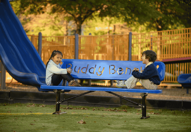 Two children sitting on a Buddy Bench in an outdoor setting, engaged in conversation or quiet reflection.