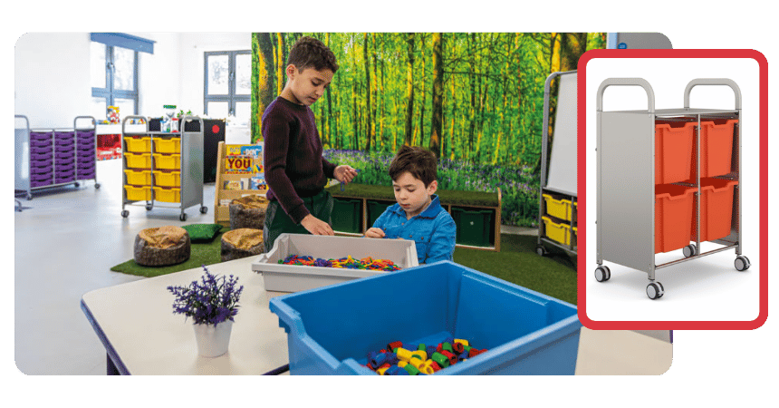 Elementary-aged children in a classroom placing items into a storage bin, engaged in organization or cleanup.