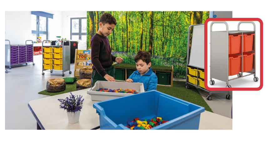 Elementary-aged children in a classroom placing items into a storage bin, engaged in organization or cleanup.