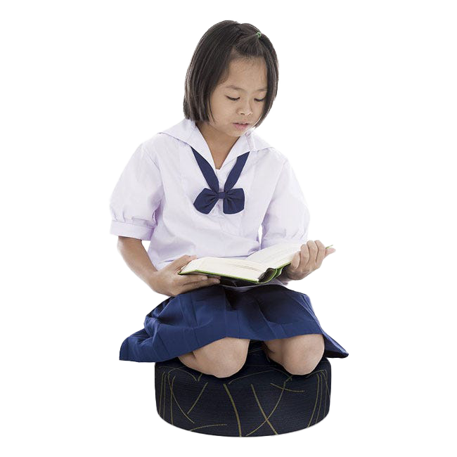 An elementary-aged child sits on a wobble stool inside her school classroom, balancing while reading a book.