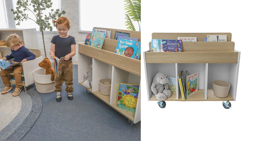 Two preschool children stand next to a wooden book display, each holding a book, inside a brightly lit preschool classroom with colorful decorations and learning materials in the background.