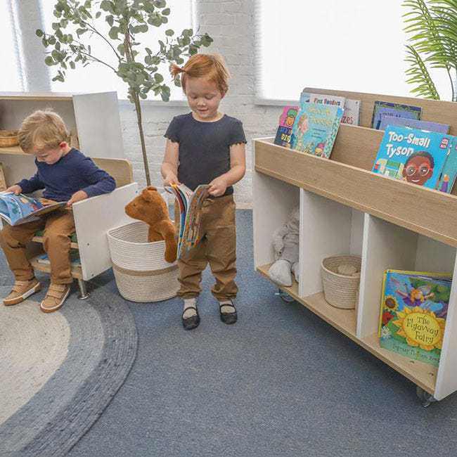 Two preschool children stand next to a wooden book display, each holding a book, inside a brightly lit preschool classroom with colorful decorations and learning materials in the background.