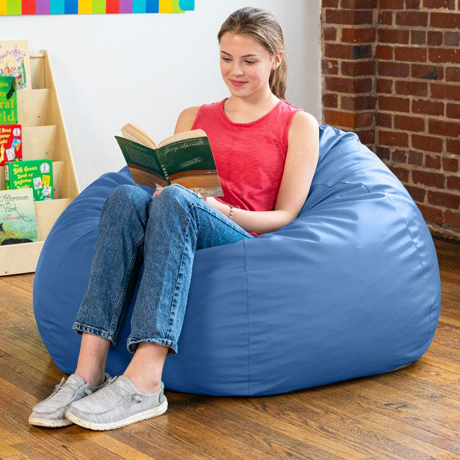 A high school student relaxes on a colorful blue beanbag inside her school library, absorbed in reading a book, with bookshelves and study areas in the background.