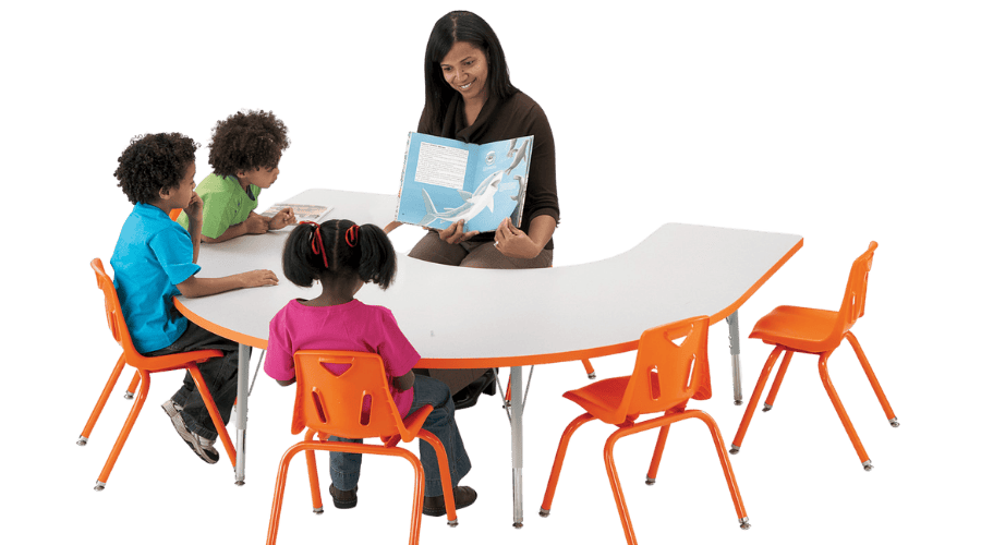 Young students gathered around a horseshoe-shaped activity table while a teacher sits in the center reading a book to them.